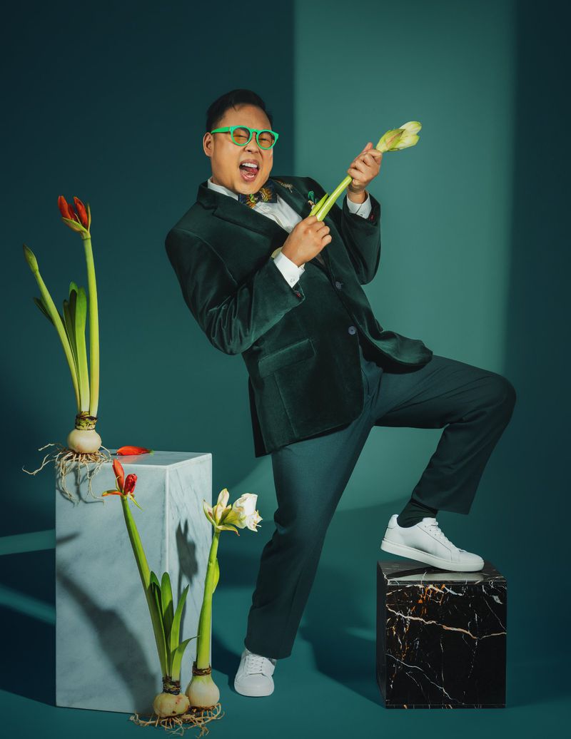 Energetic man in green suit plays flower stems as a guitar, vibrant and playful against a teal background with plants and marble cubes.