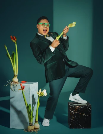 Energetic man in green suit plays flower stems as a guitar, vibrant and playful against a teal background with plants and marble cubes.