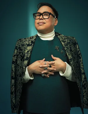 Portrait of Nico Santos wearing a patterned green blazer, white turtleneck, and statement rings against a dark teal background.