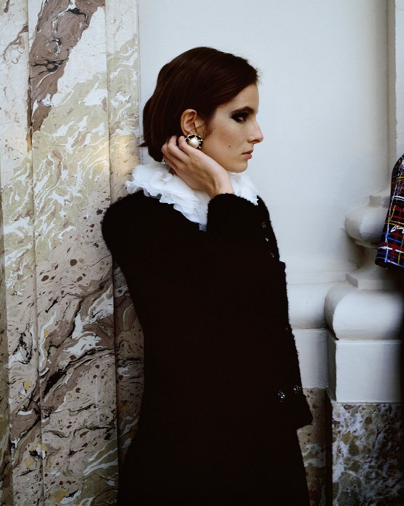 Profile view of a woman in a black jacket with a ruffled collar, pearl earrings, and dramatic eye makeup, leaning against a marble pillar.
