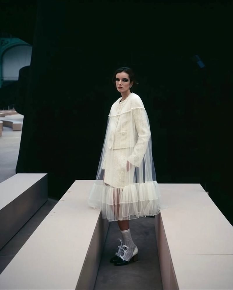 Fashion model in tweed jacket and skirt with tulle overlay, posing in a runway show setting with geometric shapes and dramatic lighting.