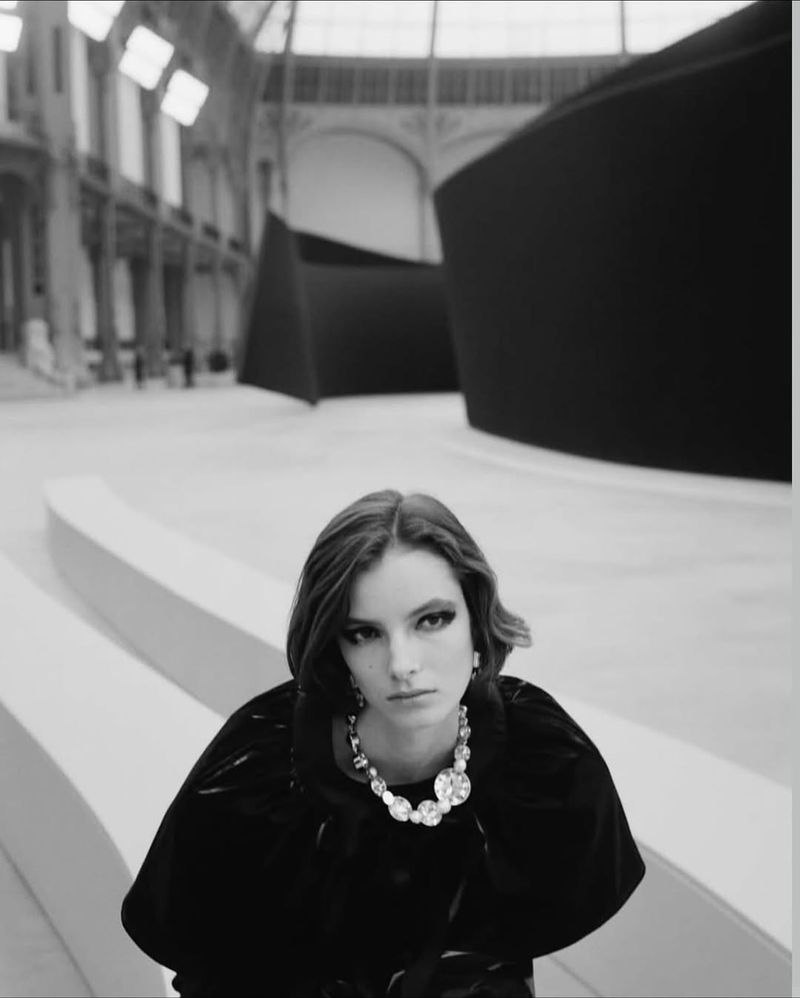 Black and white portrait of a young woman wearing a cape and statement jewelry sitting on steps in a large indoor space.