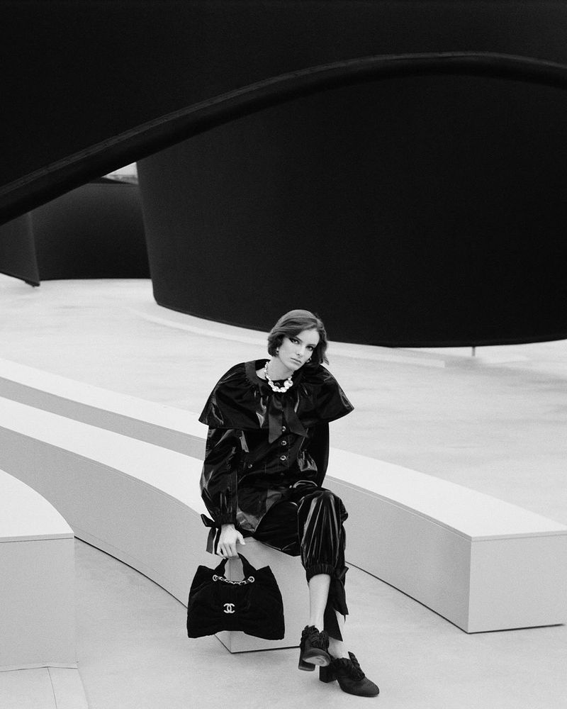 Black-and-white fashion shot of a woman in a shiny black outfit sitting on a bench, holding a handbag, indoors with modern architecture.