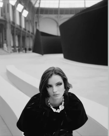 Black and white portrait of a model wearing a dark top and large crystal necklace inside a grand exhibition hall.