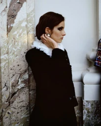 Model in black jacket with white ruffled collar adjusting pearl earring against marble pillar and white wall