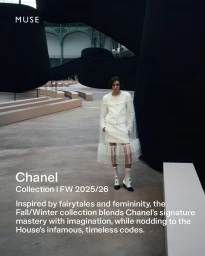 Model standing in a white tweed jacket and skirt ensemble with a sheer overlay against a large indoor architectural installation.