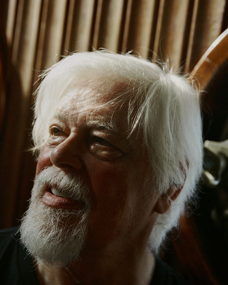 Headshot of an elderly man with white hair and beard looking up and to the left with a dark shirt and tan fabric background.