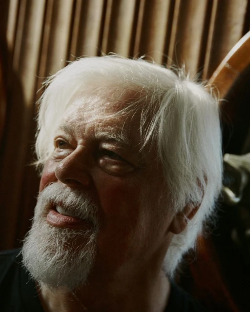 Headshot of an elderly man with white hair and beard looking up and to the left with a dark shirt and tan fabric background.