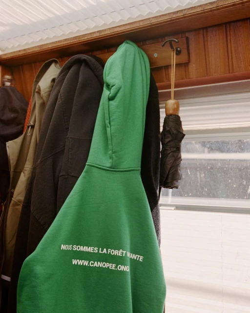 A green hooded sweatshirt hangs on a wooden coat rack, with other coats and an umbrella visible in the background against a window.