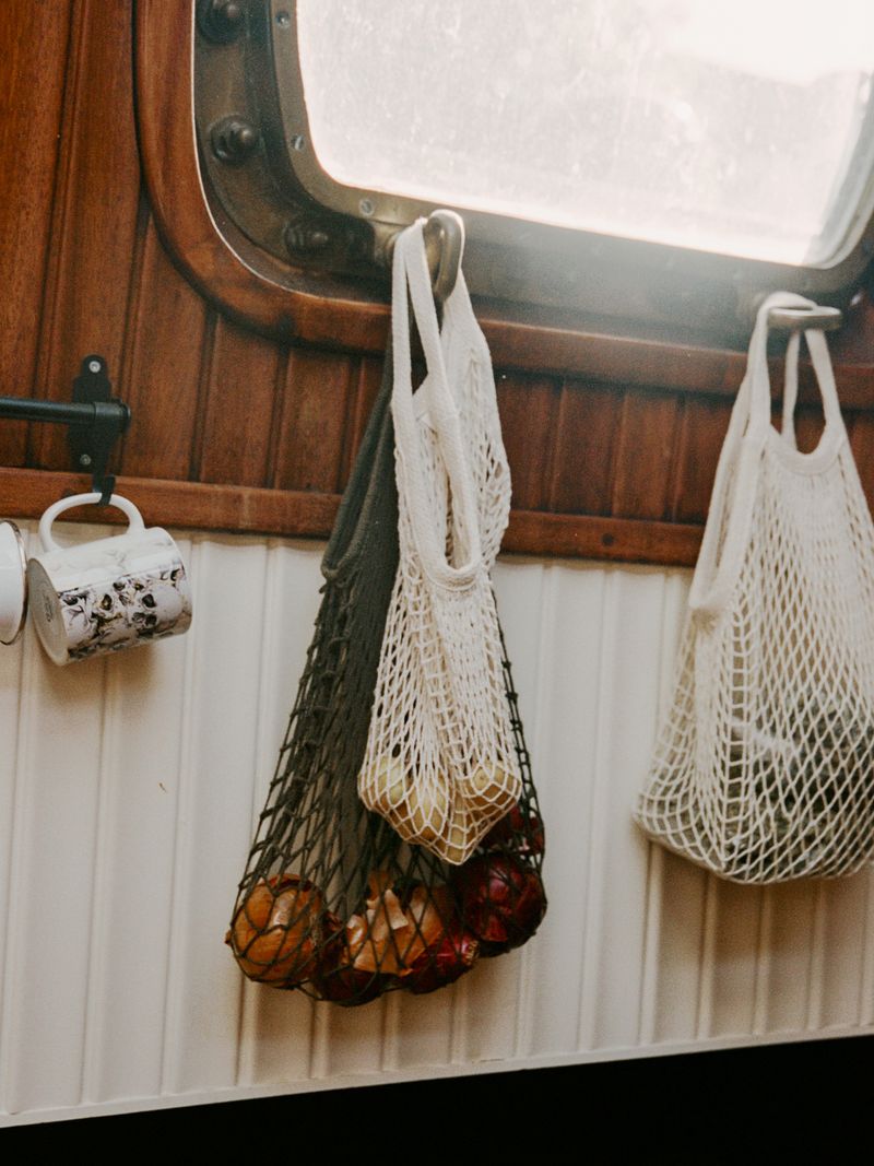 Hanging mesh bags filled with onions and other produce create a rustic kitchen storage scene with a window and paneled walls.