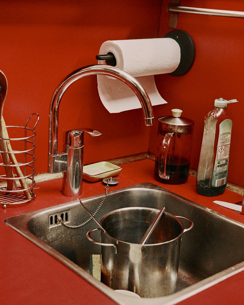 Interior shot of a kitchen sink with a metal pot, faucet, paper towels, and dish soap, set against a vibrant red wall.