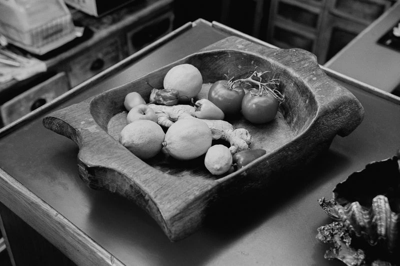 Black and white still life of a wooden bowl filled with an assortment of fruits and vegetables including lemons and tomatoes.