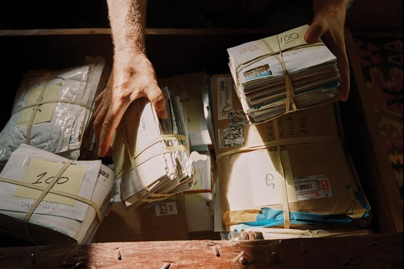 Close-up of hands placing bundles of mail, secured with rubber bands, into a wooden drawer, illuminated by sunlight streaming in.