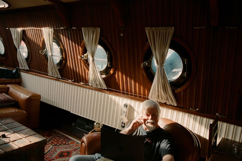 Man with laptop inside wooden yacht cabin. Porthole windows, leather furniture, nautical interior create cozy, warm atmosphere.