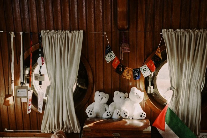 Interior shot of three white teddy bears sitting on a wooden chest, wooden wall paneling with curtains and round windows in background.