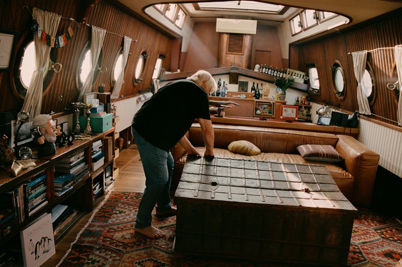 Man tending to a wooden table in a wooden-walled boat interior with books, bottles, and decorative items displayed, creating a cozy setting.