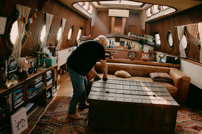 Man tending to a wooden table in a wooden-walled boat interior with books, bottles, and decorative items displayed, creating a cozy setting.