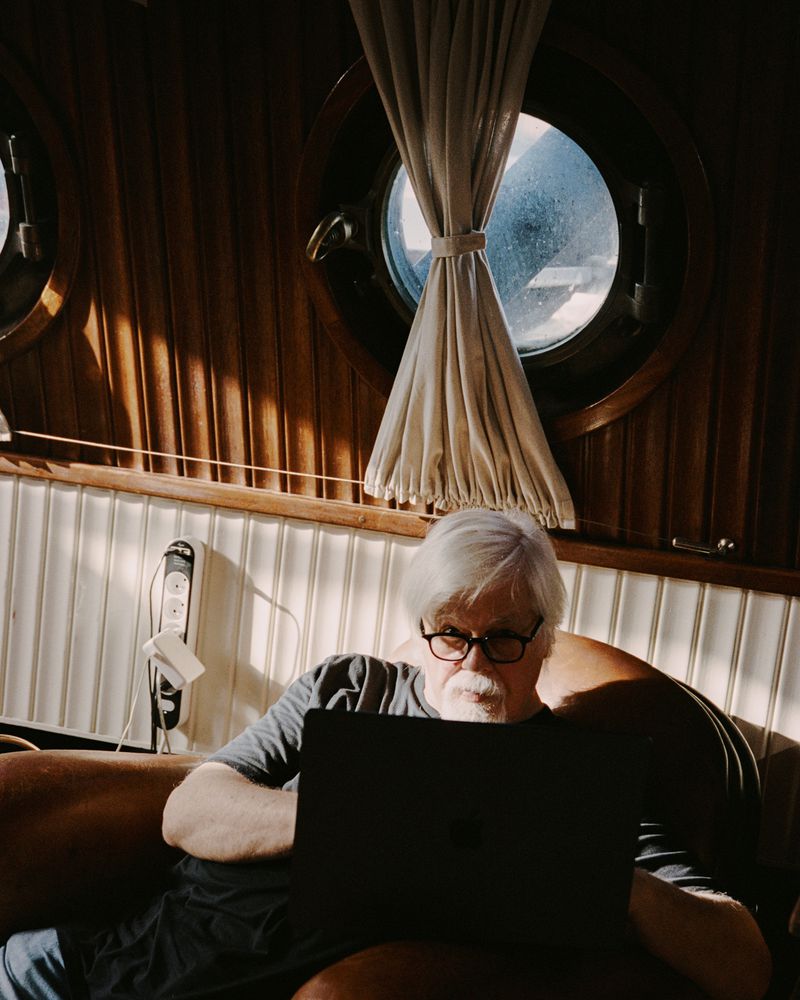 Elderly man with glasses sits indoors working on a laptop in the warm light, with a porthole window in the background.