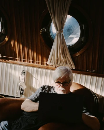 Elderly man with glasses sits indoors working on a laptop in the warm light, with a porthole window in the background.