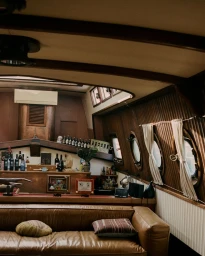Interior cabin of a ship with a leather sofa, wood paneling, circular porthole windows, and a bar counter stocked with bottles.