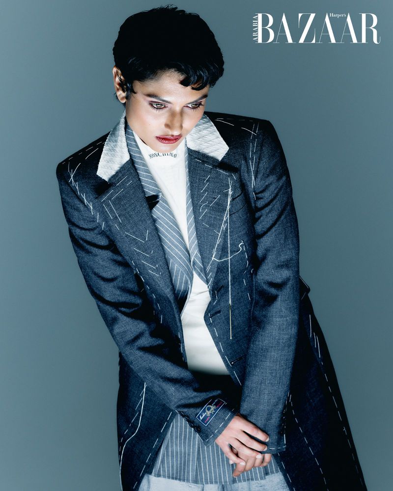 Adult female fashion model poses in a jacket with tailor markings for Harper's Bazaar, shot in a photo studio against a grey backdrop.