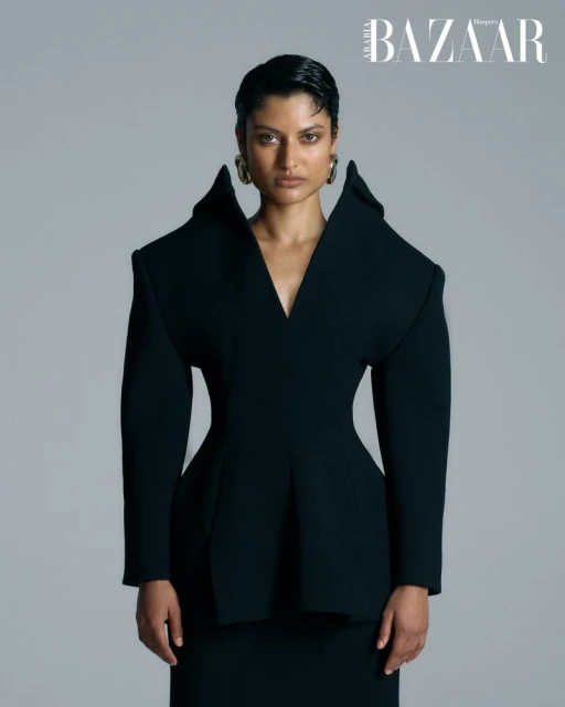 Fashion editorial shot of an adult woman wearing a dark, structured couture jacket and skirt with statement earrings against a grey backdrop.