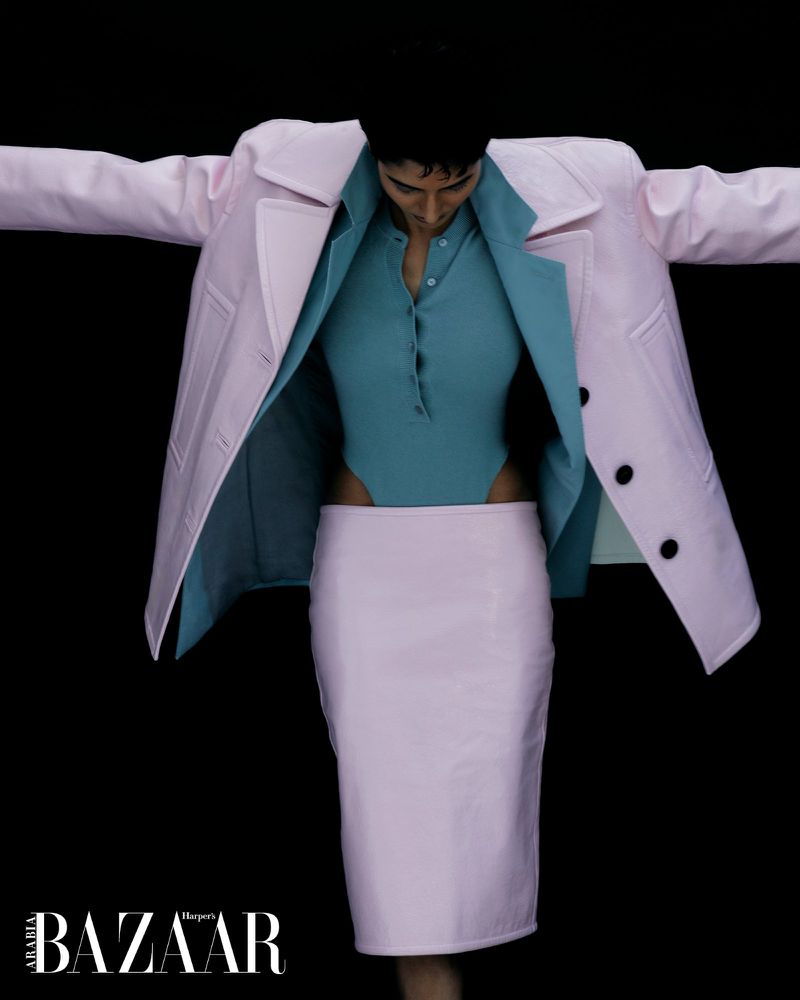 Fashion shot featuring a model in a pink coat and skirt, accessorized with a blue bodysuit, against a stark black background.