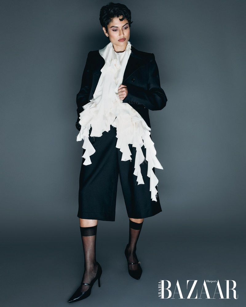 Fashion editorial shot: Woman in black blazer, culottes, Mary Jane heels, and ruffle scarf poses against a gray backdrop.