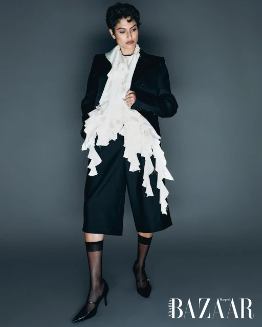 Fashion editorial shot: Woman in black blazer, culottes, Mary Jane heels, and ruffle scarf poses against a gray backdrop.