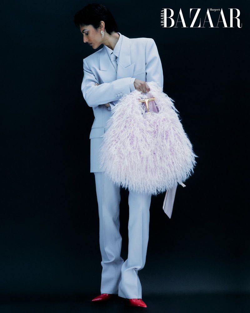 Full-length studio shot of a model in a light blue suit holding a fuzzy lavender handbag against a dark background, fashion editorial.