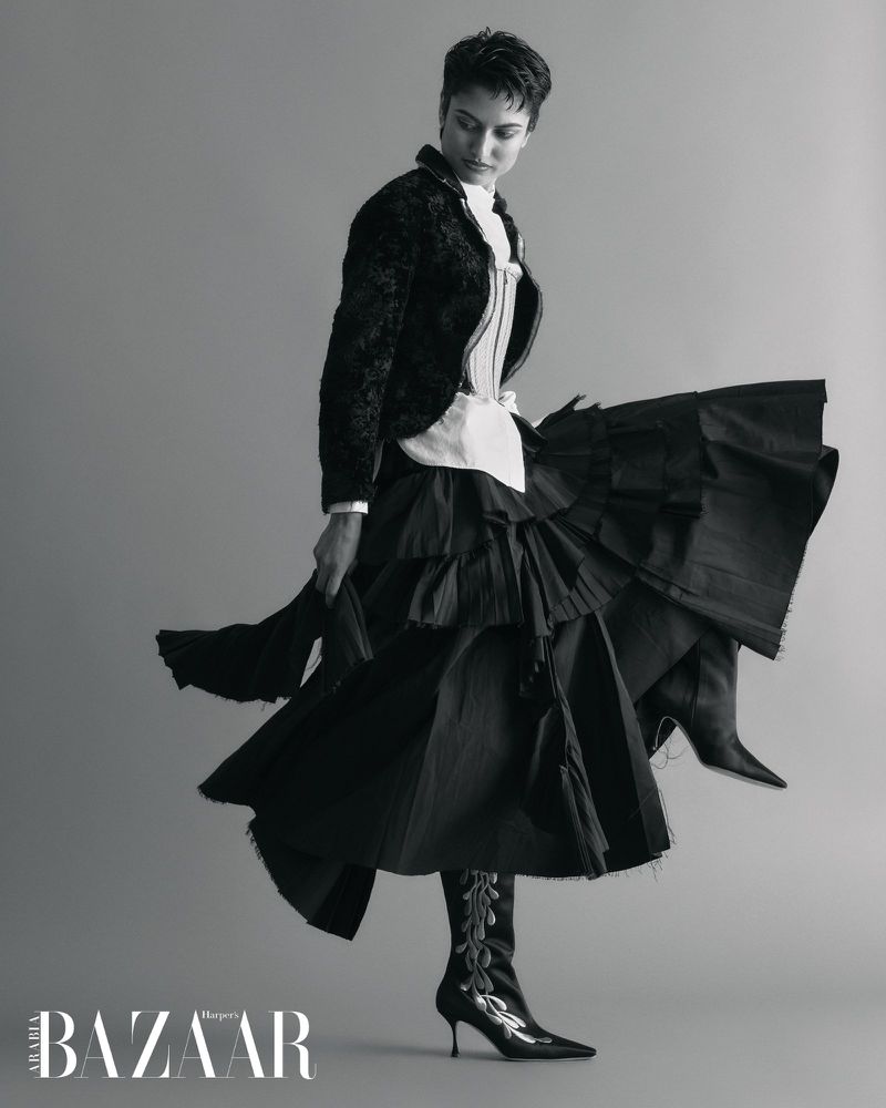 Model in dark textured jacket and voluminous flared skirt posing against a seamless grey backdrop for Harper's Bazaar Arabia