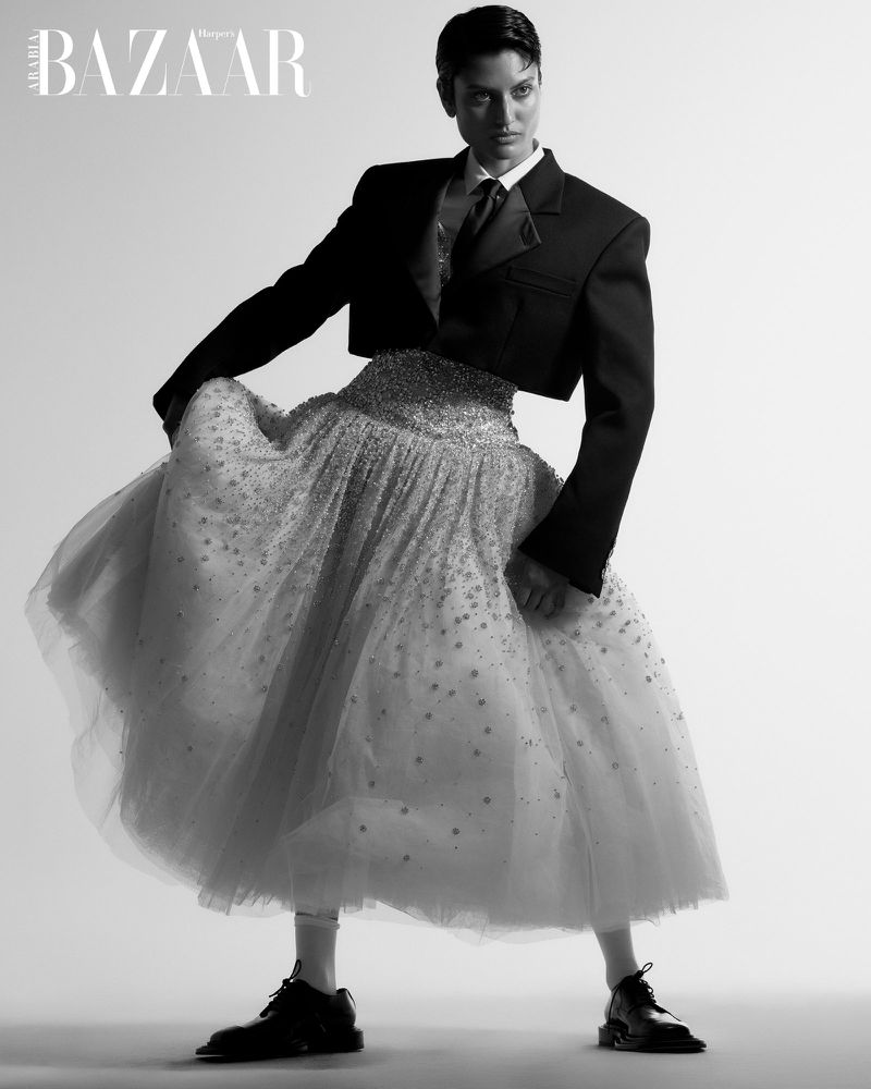 Black and white portrait of a woman in a cropped blazer and tulle skirt, standing in a light, neutral studio environment with soft lighting.