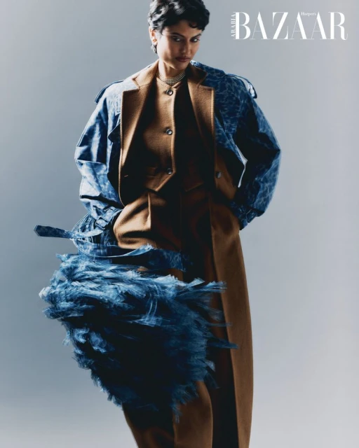 Fashion editorial shot of a woman in a tan suit and denim jacket with feathered embellishments against a light gray backdrop.