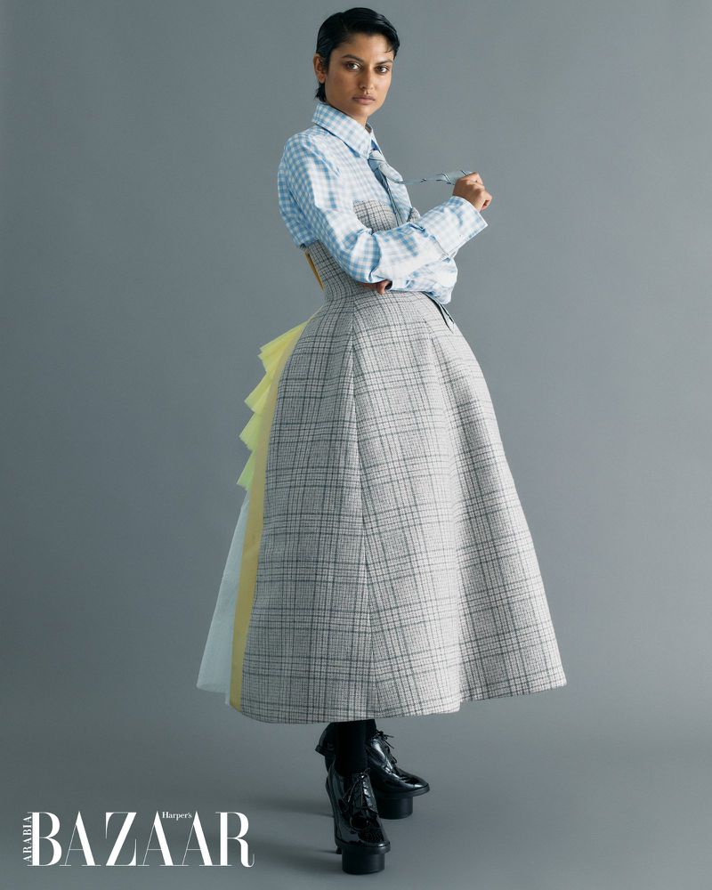 Fashion editorial shot of a woman in a plaid dress with black platform shoes against a grey background. The model is indoors.