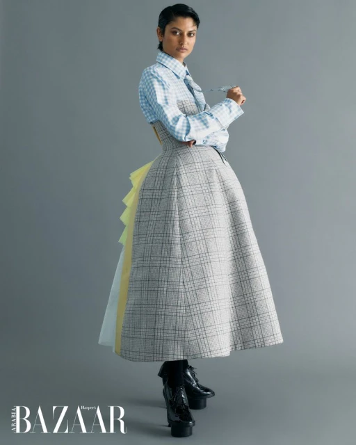 Fashion editorial shot of a woman in a plaid dress with black platform shoes against a grey background. The model is indoors.
