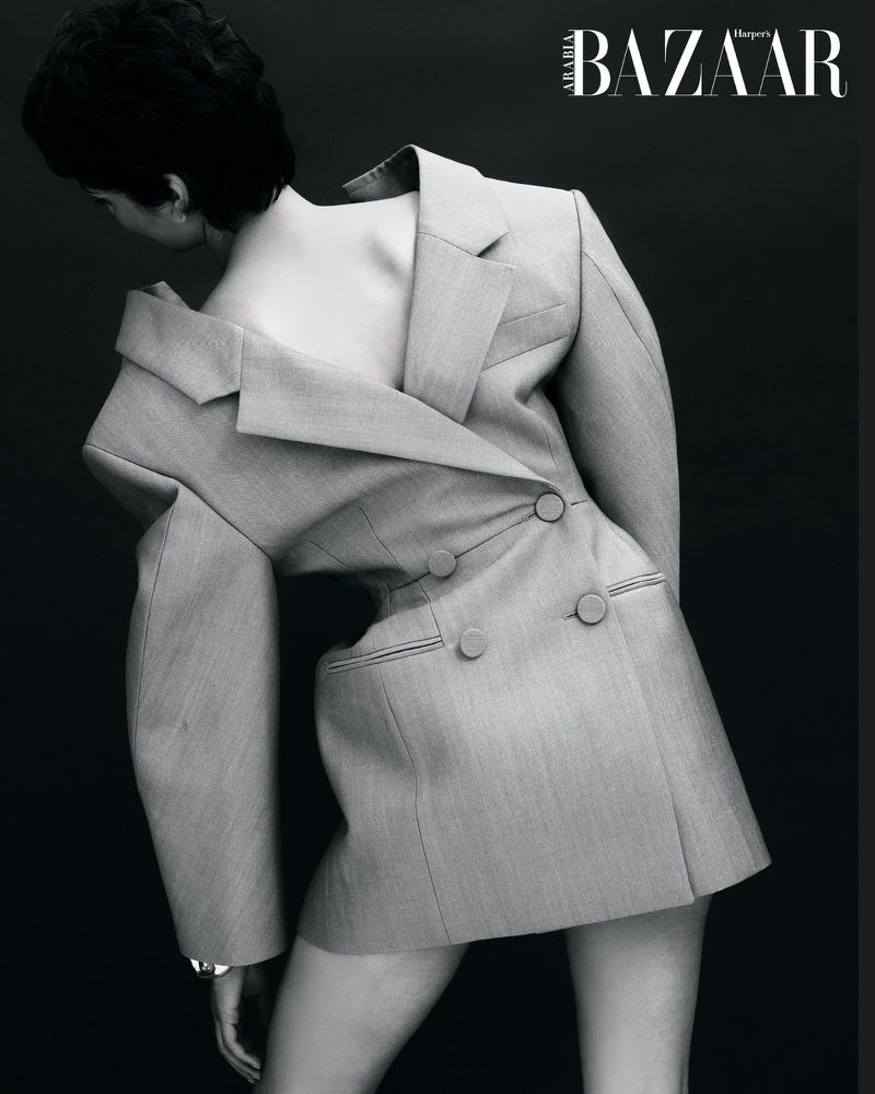 Fashion editorial shot of an adult woman wearing an oversized grey blazer, posing in front of a dark grey background, black and white.
