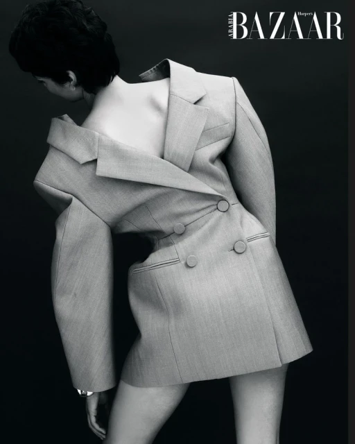 Fashion editorial shot of an adult woman wearing an oversized grey blazer, posing in front of a dark grey background, black and white.