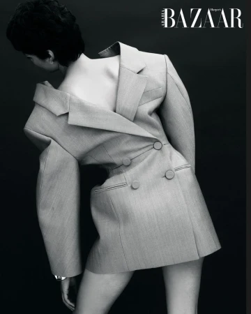 Fashion editorial shot of an adult woman wearing an oversized grey blazer, posing in front of a dark grey background, black and white.