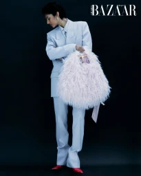 Model in a pale blue suit holding a fluffy pink feathered bag against a dark background for Harper's Bazaar Arabia