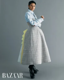 Model Bejal Desai in a blue gingham shirt and a structured grey plaid skirt with layered back ruffles against a grey studio backdrop.