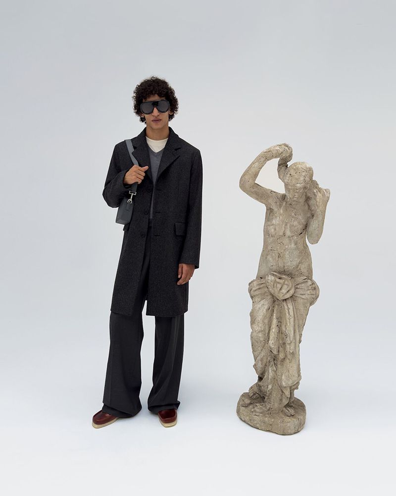 A male fashion model poses in a dark coat and pants next to a stone sculpture against a plain white background in a studio setting.