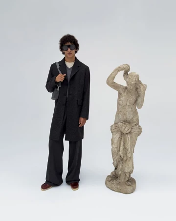 A male fashion model poses in a dark coat and pants next to a stone sculpture against a plain white background in a studio setting.