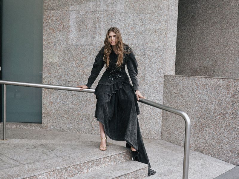 Stylish adult female poses on steps in long tiered dark dress and heeled sandals against a textured neutral urban backdrop.