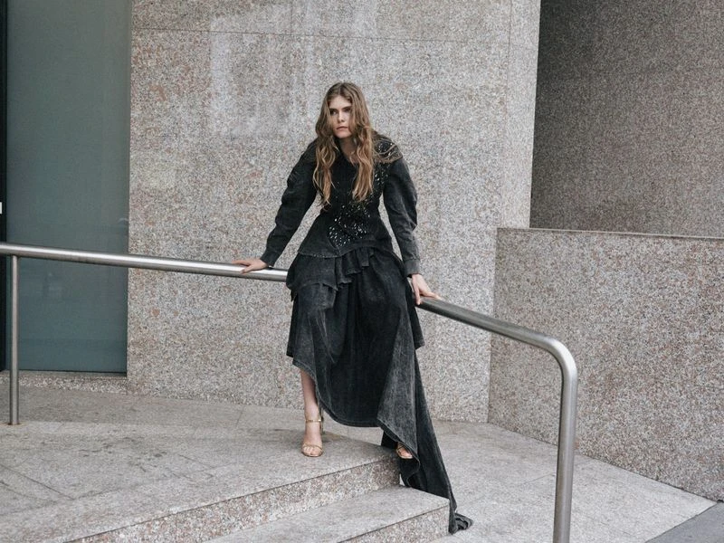 Stylish adult female poses on steps in long tiered dark dress and heeled sandals against a textured neutral urban backdrop.