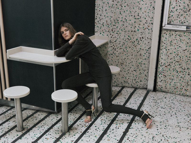 Fashion editorial shot of a young adult woman in black clothing seated at a white table in a speckled interior with white stools.