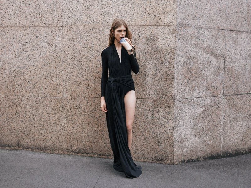 A slender adult is outdoors against a textured wall wearing a black dress and sipping from a takeaway cup. The person faces forward.