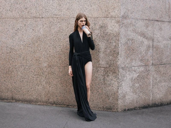 A slender adult is outdoors against a textured wall wearing a black dress and sipping from a takeaway cup. The person faces forward.
