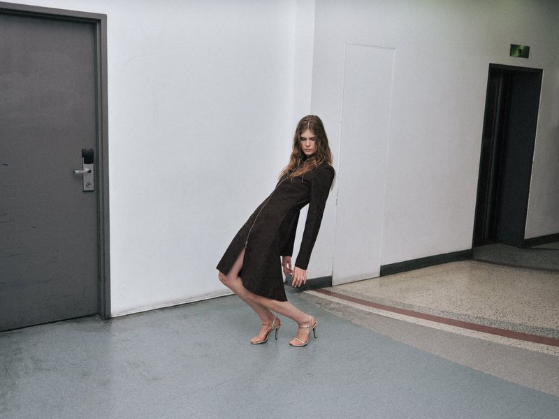 Fashion shot: A slender woman in a long brown dress and high heels leans against a wall in an indoor corridor with gray doors.
