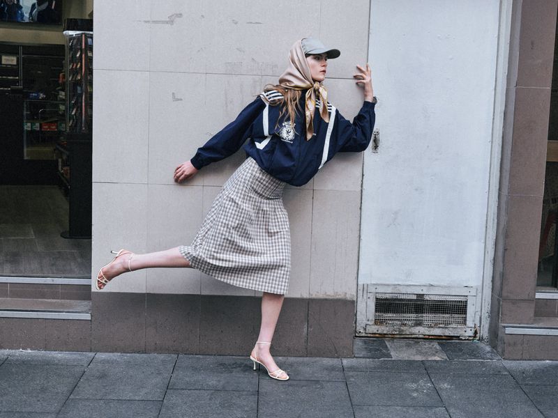 A woman in a navy jacket, plaid skirt, and headscarf playfully leans against a wall on a city sidewalk, posing for a fashion shot.