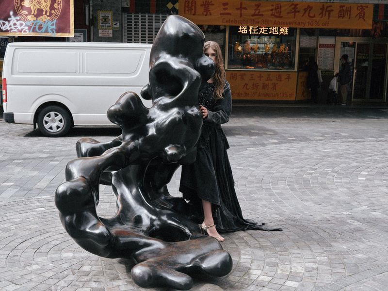 Woman poses in a flowing black dress beside a dark, abstract sculpture on a city street with a white van and storefronts in the background.